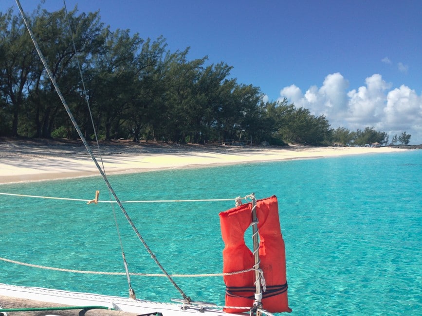 Image shows an orange lifejacket hanging on a sailboat's rail. Behind the lifejacket, there is blue water, a sandy beach and a thicket of green trees.