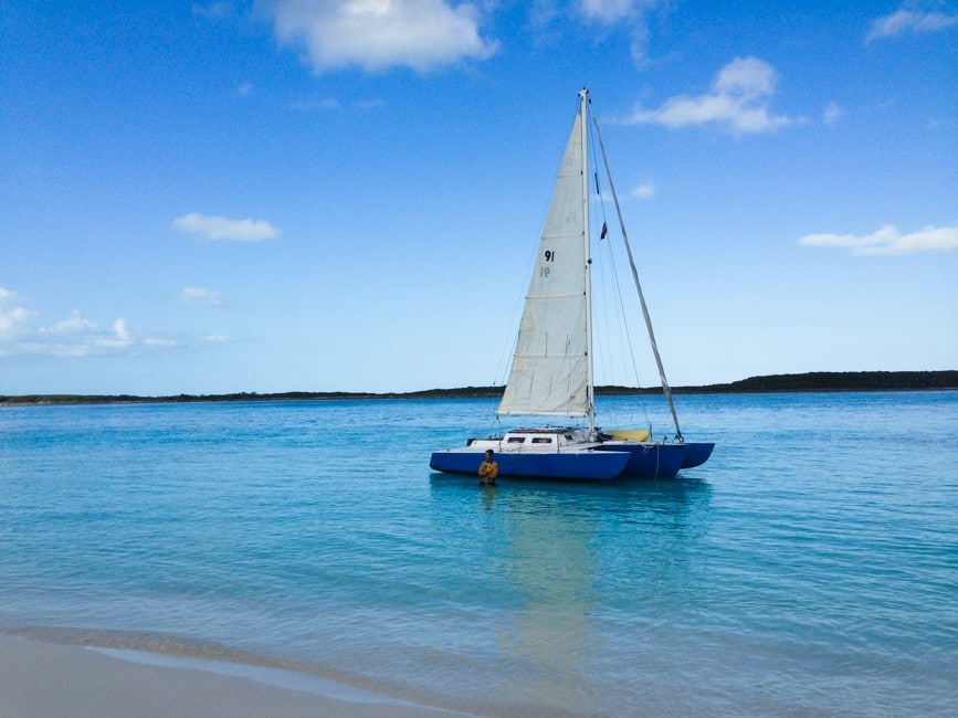 The image shows a picture of a sailboat anchored in the Bahamas. The boat is a small catamaran, painted blue, and the sails are up.