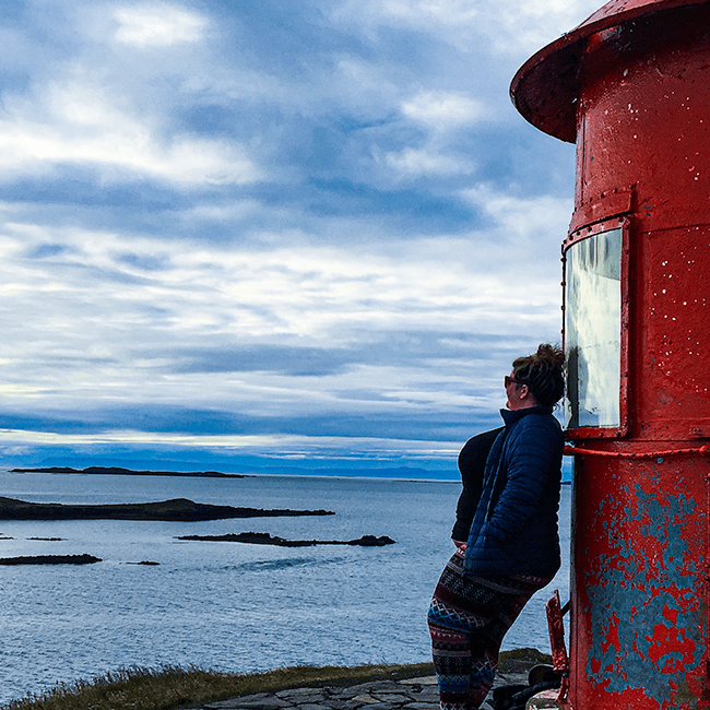 Jezebel is standing against a red lighthouse, looking out at the ocean. She is wearing hiking gear and a blue coat. The sky is scattered with clouds.