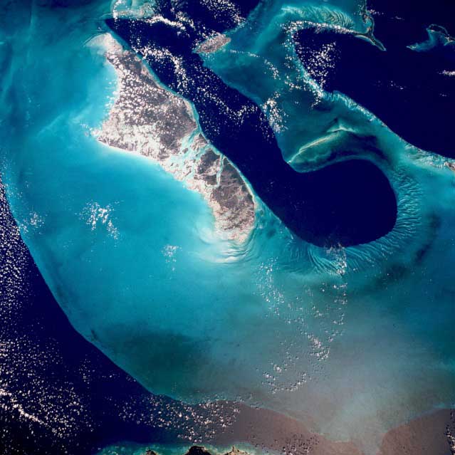 Image shows an overhead view of the Tongue of the Ocean, the depest water in the Bahamas. Light blue water surrounds a long channel of black-blue water between two islands.