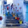 Image shows a stairwell in the Moroccan city of Chefchaouen. The medina is painted blue, with doors and stairs painted different shaes of blue. Along the stairwell, brightly coloured flowerpots hang against walls.