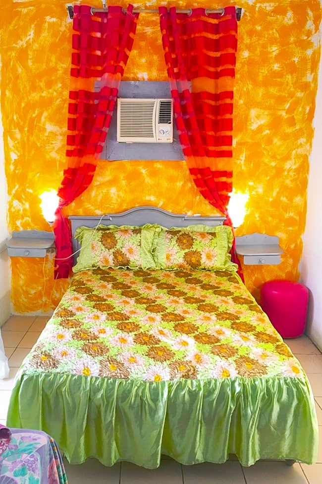 The photo features a full-sized bed with a bright orange and yellow bedspread with a green bedskirt. The wall is painted bright orange and above the bed there are red curtains.