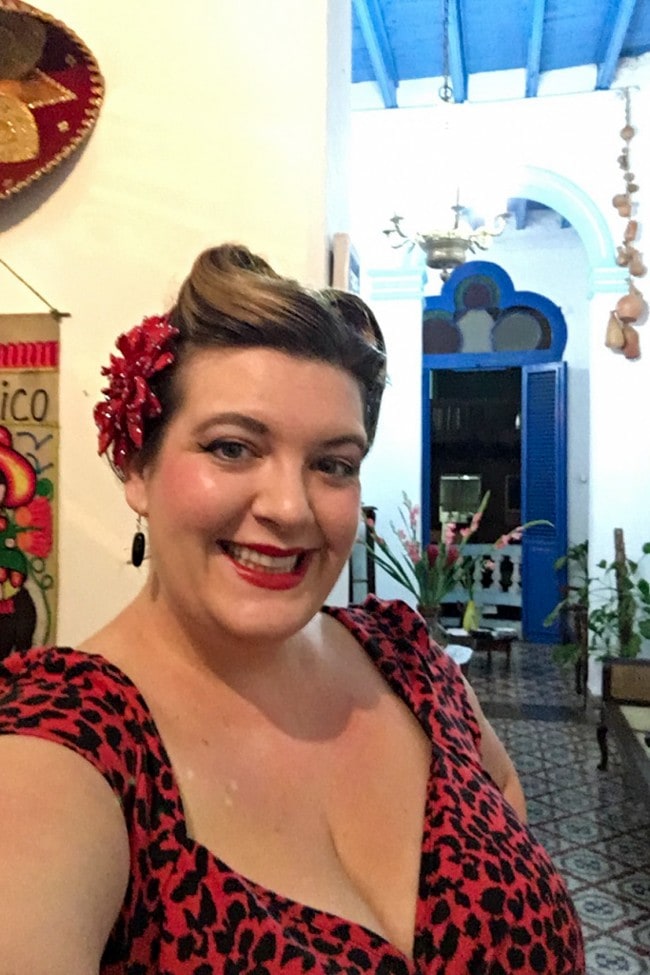 Jezebel stands in a brightly painted hallway, smilign at the camera. She is wearing a red leopard print dress and a large red flower in her hair.