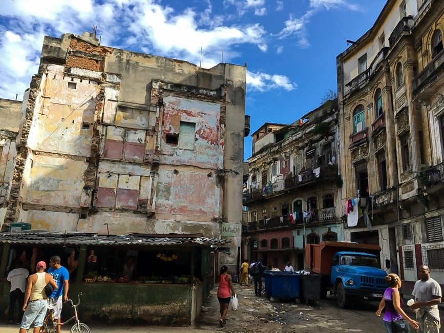 The photo shows a narrow street in havana. The sky is blue above the buidlings, and the buildings are in poor repair.