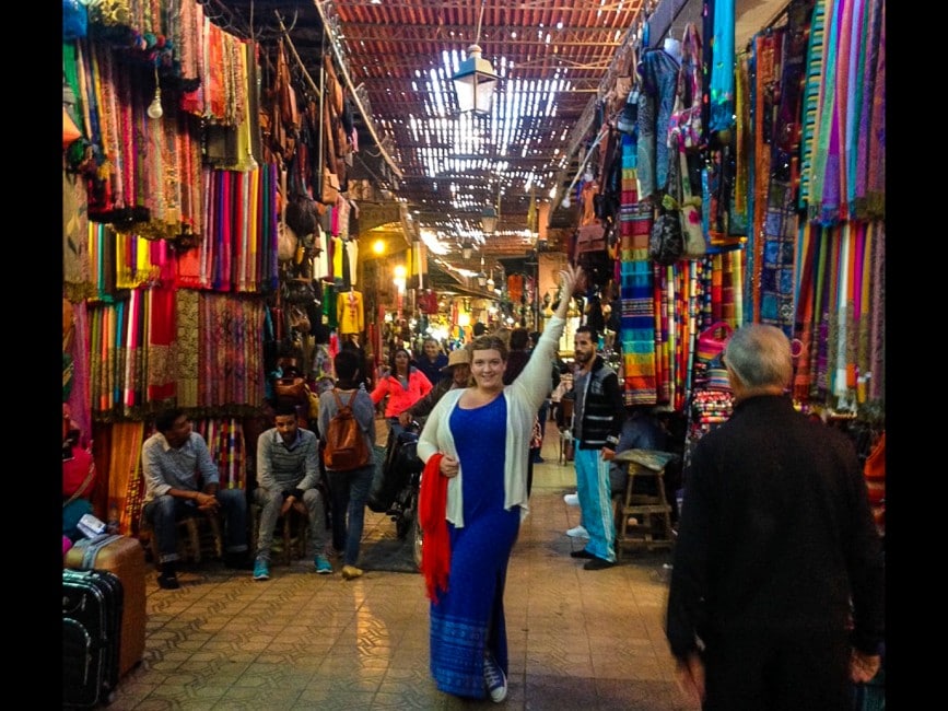 Image shows Jezebl striking a showgirl pose in the crowded market of the Marrakech Medina. Fabrics and items for sale are piled to the ceiling in brightly coloured displays.